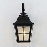 A black metal outdoor wall lantern with a clear glass enclosure, mounted on a white wall, and featuring an exposed glowing Edison-style bulb inside.