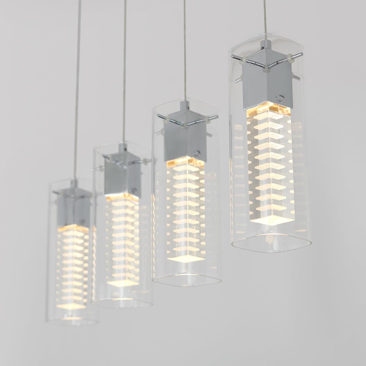 Four modern pendant lights with clear cylindrical glass shades and metallic fixtures hang in a row, each glowing with a soft white light against a plain, light-colored background.