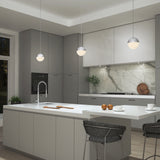 Modern kitchen with gray cabinets, a white island with sink, three spherical pendant lights, marble backsplash, and bar stools. Cutting boards and apples rest on the island; plants add greenery to the countertop.
