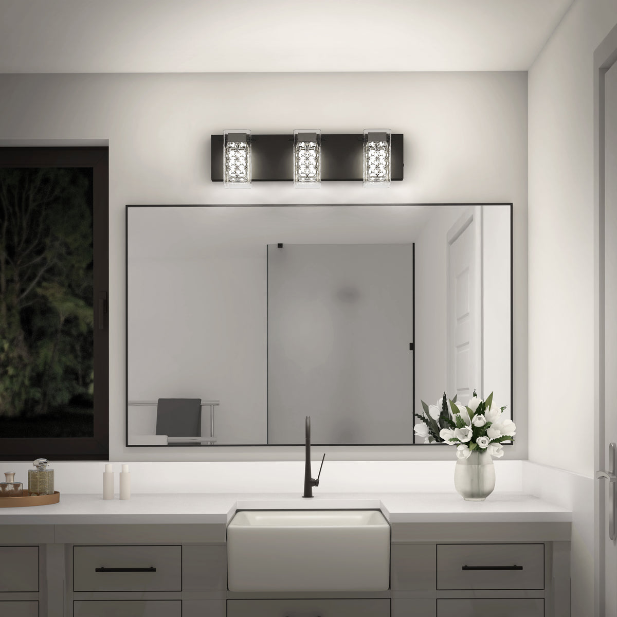 A modern bathroom with a large mirror, white countertop, rectangular sink, black faucet, a vase of white flowers, and a three-light fixture above the mirror. A window shows a dark, tree-filled view outside.