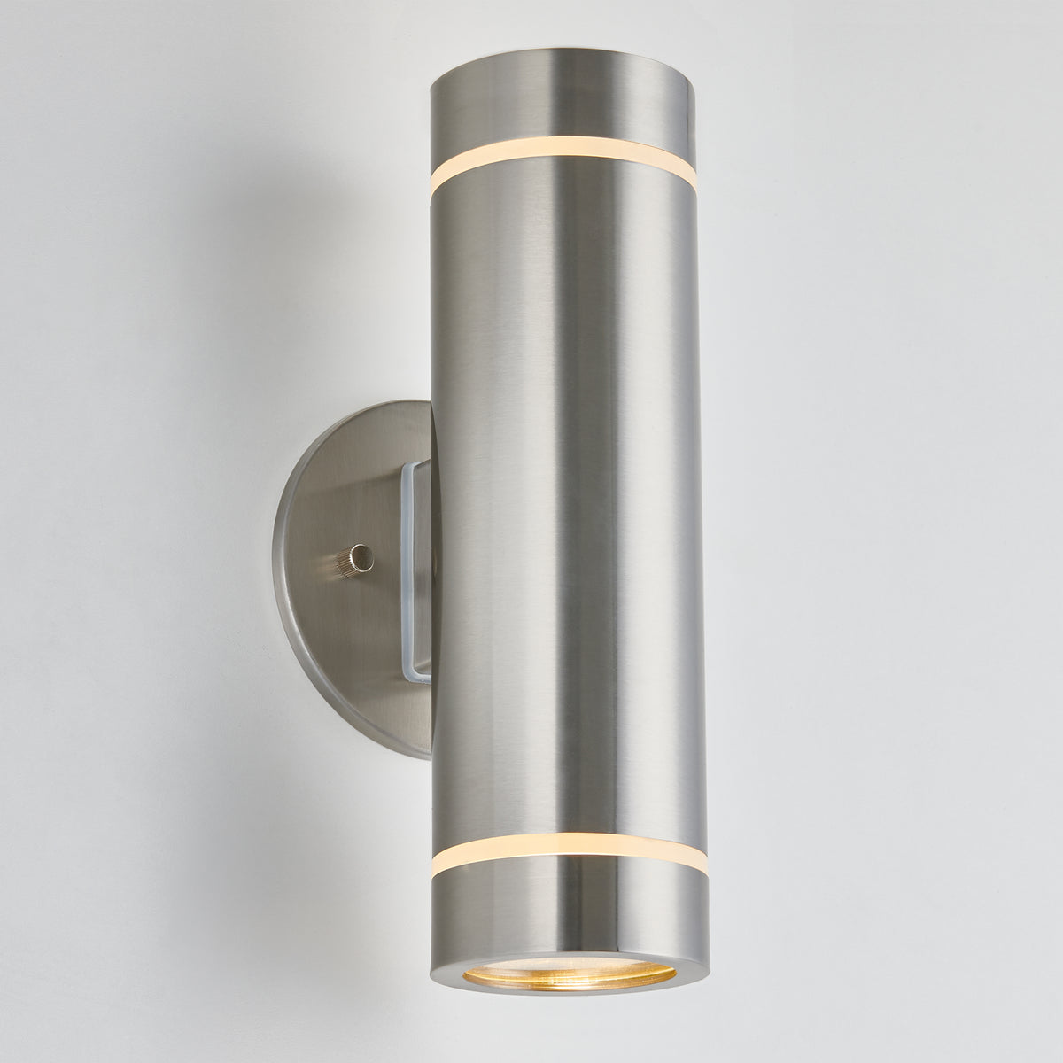The Artika C7 indoor-outdoor wall light features a modern cylindrical brushed metal design, emitting soft white light from both ends and mounting on a round plate—ideal for versatile indoor or outdoor lighting.