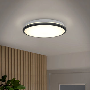The Artika Vera 13 in. Modern LED Flush Mount ceiling light shines in a wood-paneled room with a door and leafy plant.