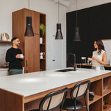A man and woman stand in a modern kitchen, each holding a mug and smiling at each other across a marble-topped island with black chairs. Wooden cabinets, pendant lights, and kitchen decor are visible.