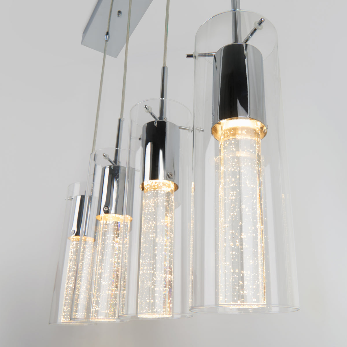 Four modern pendant lights with cylindrical glass shades and chrome accents hang in a row. The inner cylinders are illuminated, creating a sparkling effect with tiny lights inside.