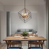 A modern dining room with a wooden table and six chairs, a geometric pendant light above, and a small plant centerpiece. In the background, there is a gray sofa with throw pillows and a blanket in a cozy living area.