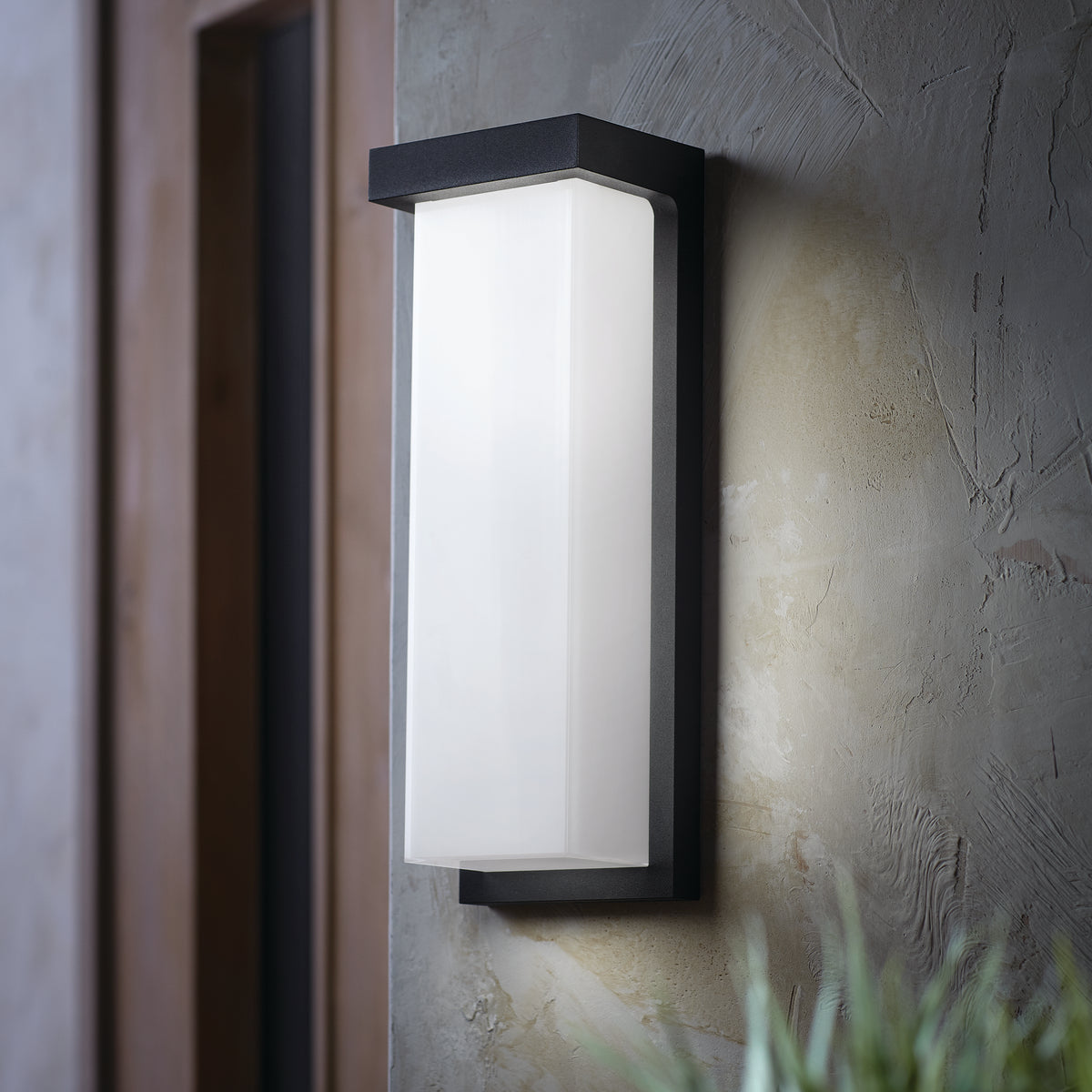 A modern rectangular outdoor wall sconce with a black frame and frosted glass is mounted on a textured gray wall near a doorway, with some green foliage visible at the bottom right.