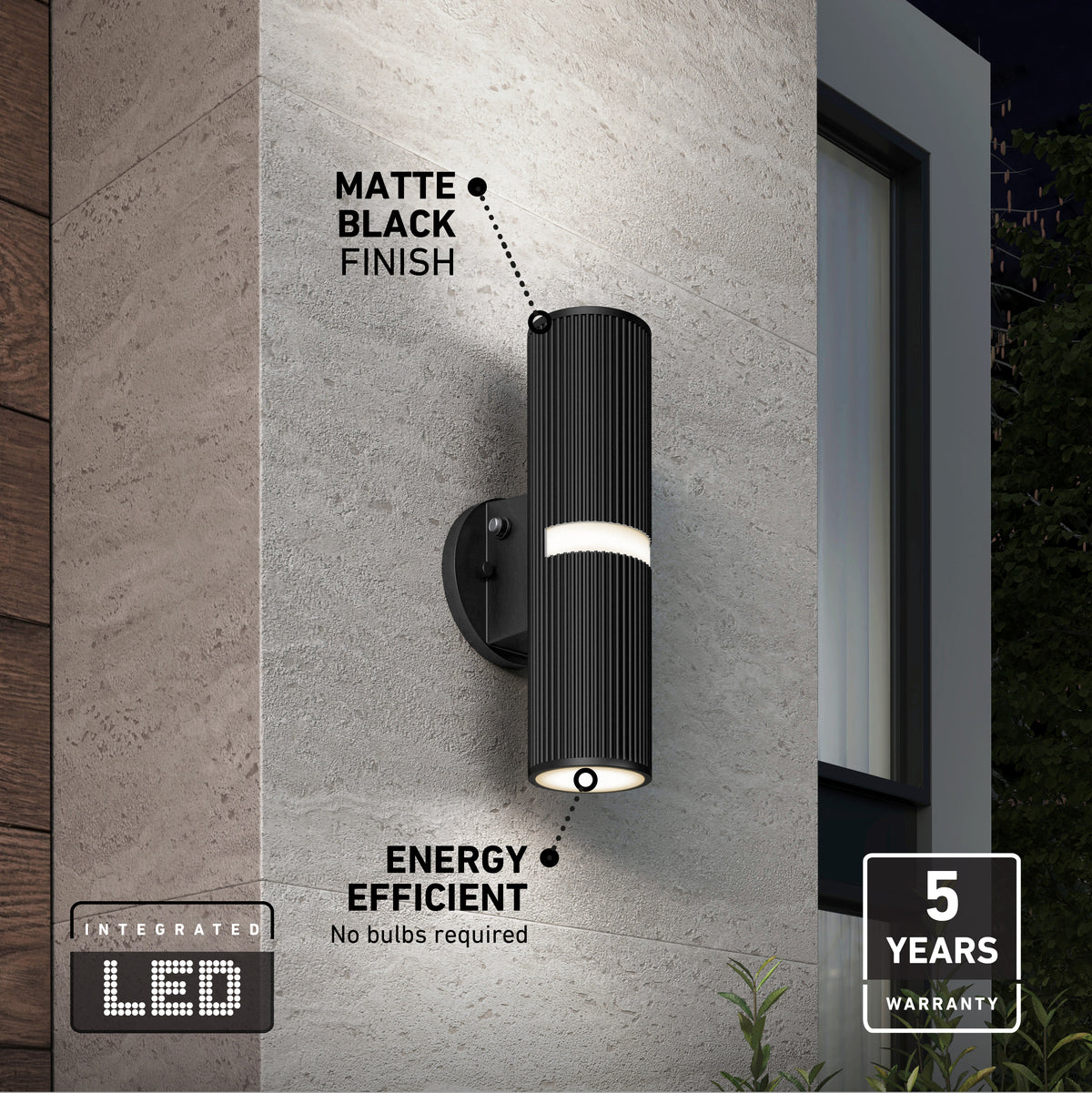 Modern black cylindrical LED wall light with matte finish mounted on an exterior wall, labeled as energy efficient (no bulbs required) and includes a 5-year warranty. Nighttime setting near a window and plants.