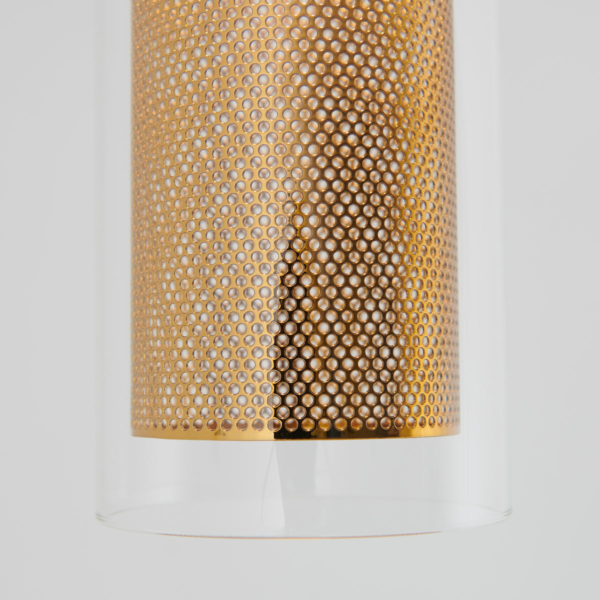 Close-up of a cylindrical object with a perforated gold metal pattern inside a clear glass tube, set against a plain light gray background.