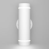 A modern cylindrical wall light fixture with a matte white finish, mounted vertically on a round wall plate, emitting light from narrow openings at the top and bottom. The background is plain and light gray.