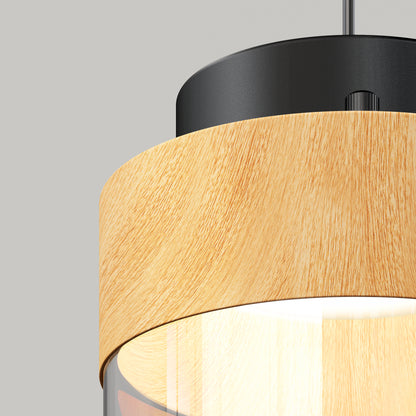A close-up of the Artika Hanson 1-light LED pendant 5 CCT, showcasing a black top, cylindrical light wood accent, and clear glass shade with soft light, set against a plain gray background.