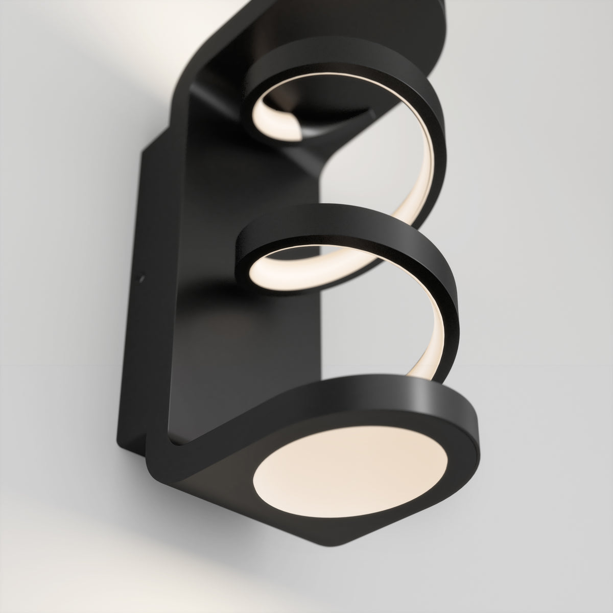 A modern black wall sconce with circular LED light elements, mounted on a light-colored wall. The design features two open rings and a round base, emitting soft, white light.
