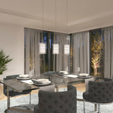 A modern dining room with a glass table set for four, surrounded by gray upholstered chairs, under three pendant lights. Large windows with sheer curtains reveal a landscaped garden with illuminated trees.