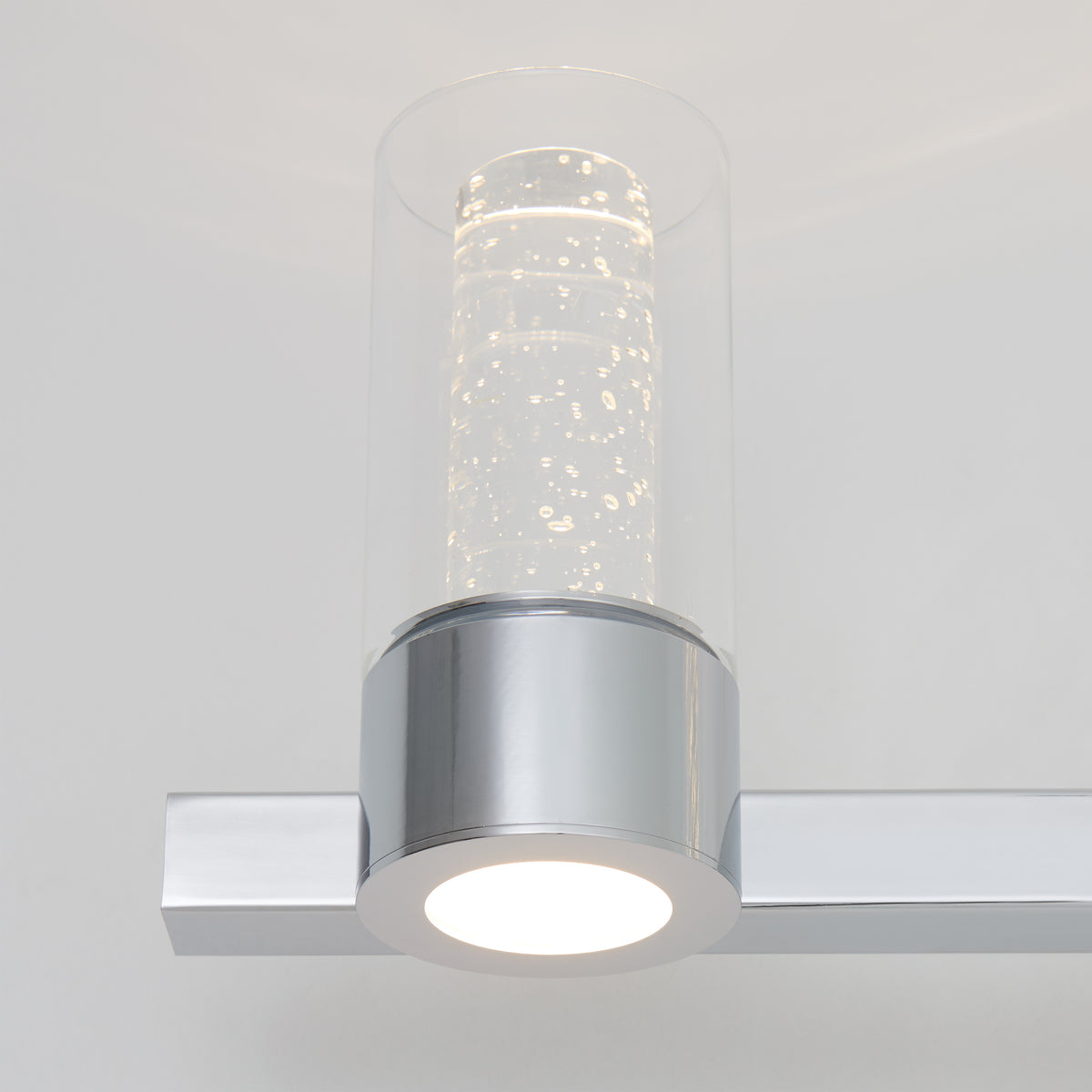 A modern wall-mounted light fixture with a cylindrical glass shade containing bubbles and a metallic silver base, emitting a soft white glow.