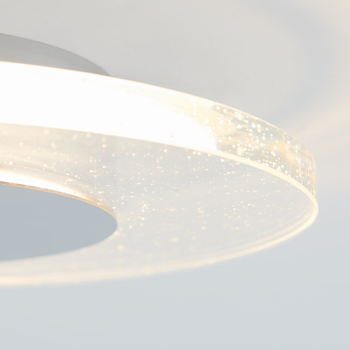Close-up of the Artika Essence Disk LED flush mount, a modern ceiling light with a translucent ring and bubble glass detail that emits a soft glow. The background is light and softly blurred.