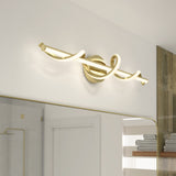 Modern bathroom vanity light with a gold, twisted LED bar design mounted above a large mirror. The fixture emits soft, white light, and towels are visible on shelves in the background.