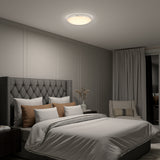 A modern bedroom with a tufted gray headboard, layered pillows, neutral bedding, a nightstand with a vase, a cushioned chair, floor-length curtains, and a round ceiling light illuminating the space.