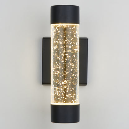 The Artika Bubble Element LED indoor-outdoor light features a cylindrical glass body filled with small bubbles, illuminated from both ends, and mounted on a black rectangular base against a light-colored wall.