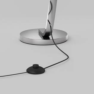 A close-up of the Artika Arc LED Floor Lamp’s chrome base with a power cord connected to a round black foot switch, resting on a smooth light gray surface.