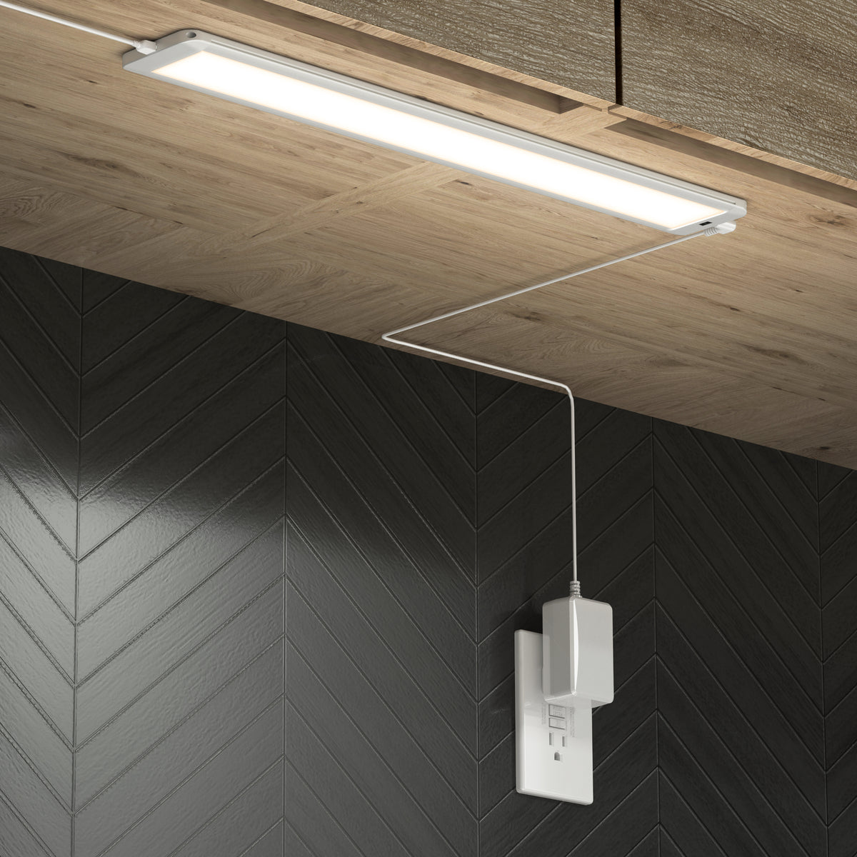 A slim LED light fixture is mounted under a wooden cabinet, with its power cord plugged into a white outlet on a black chevron-patterned wall.