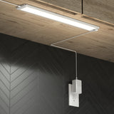 A slim LED light fixture is mounted under a wooden cabinet, with its power cord plugged into a white outlet on a black chevron-patterned wall.
