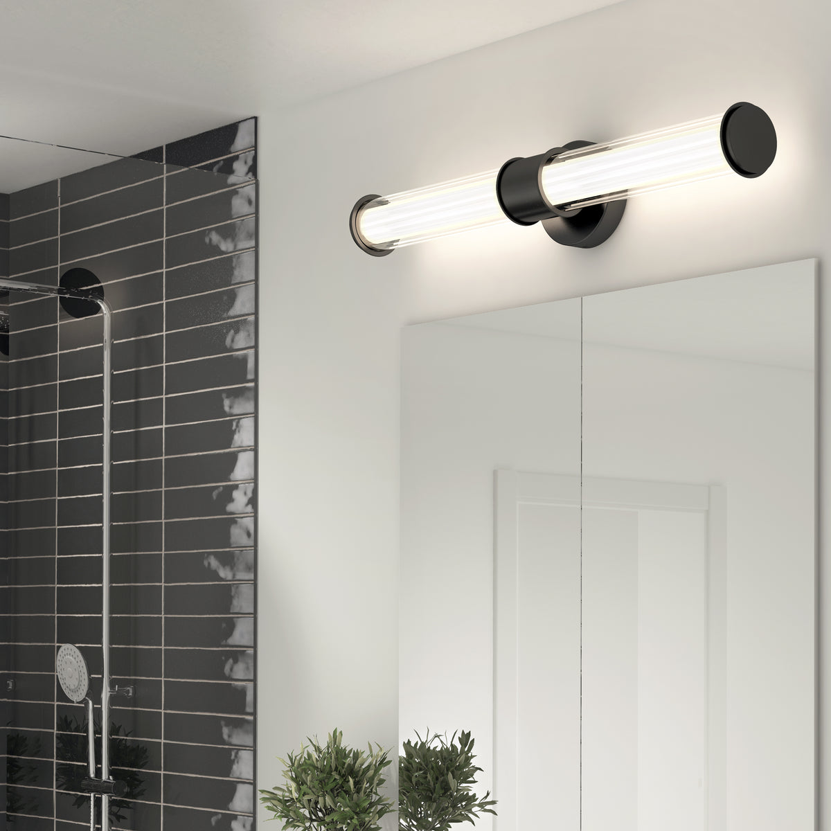 A modern bathroom light fixture with two tubular bulbs mounted horizontally above a large mirror, next to a tiled shower area and a small green plant on the counter.