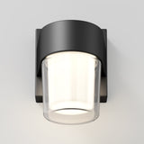 A modern wall-mounted light fixture with a matte black square base and a cylindrical frosted glass shade, emitting a soft, warm light against a plain white background.