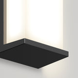 Close-up of a modern wall-mounted light fixture with a rectangular black metal base and a frosted glass shade, glowing softly against a light-colored wall.