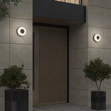 A modern entrance with a wooden door, light stone tile walls, two circular wall lights on either side, and two potted plants in black containers flanking the doorway.