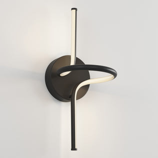 The Artika Swirl LED Sconce features a circular black base and a single curved LED light tube in an abstract design, beautifully mounted on a plain white wall.