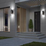Modern house entrance at dusk with a black door labeled “05,” two wall lights, a potted evergreen shrub on the steps, large windows with sheer curtains, and a decorative wood slat panel beside the door.
