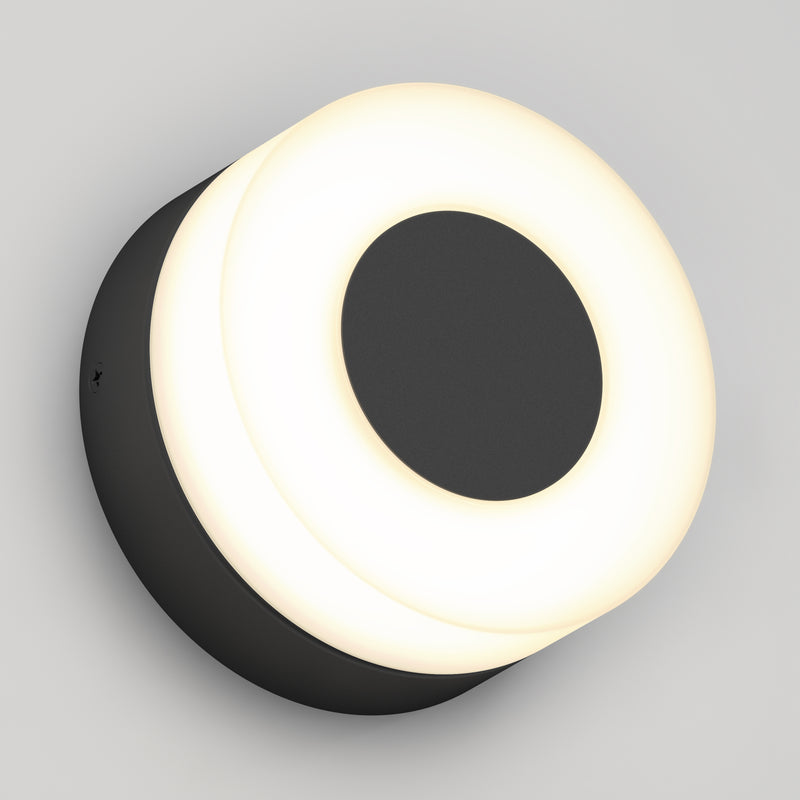 The Artika Ludwig modern LED indoor-outdoor wall light 5CCT features a round black central circle and outer ring, emitting a soft, even white glow, and is shown mounted on a light gray wall.