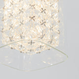 A close-up of a modern light fixture with a geometric, crystal-like pattern, illuminated from within and reflecting softly on a curved glass surface below. The background is a plain, light gray.