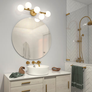 Modern bathroom with a round mirror above a white vanity, gold faucet and fixtures, vessel sink, globe light, green towels, the Artika Bloom 27-in 3-Light Modern LED Vanity Light for elegant lighting, and a glass shower with gold accents and herringbone tiles.