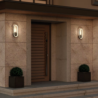 Modern front entrance with a wooden door, stone walls, steps, and two Artika Aiden modern LED indoor-outdoor wall lights on either side. Black square planters with potted plants flank the doorway.