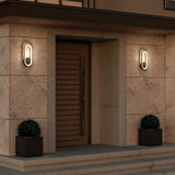 Modern house entrance with a wooden slatted door, two oval wall lights on either side, beige stone walls, and two potted round shrubs in square planters flanking the doorway.