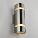 A modern cylindrical wall sconce light with a brushed metal finish, mounted on a rectangular back plate, emitting soft light from the top and bottom openings against a plain light gray wall.