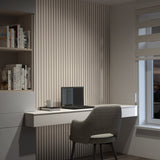A modern home office with a gray upholstered chair, a white floating desk with a laptop and mug, vertical slat wall paneling, built-in shelves with books, and a window with layered blinds.