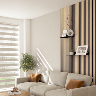 A modern living room with a beige sofa, brown cushions, a potted plant, and soundproof wall panels. Two Artika Sonokit shelves and hooks for Sonolok and Sonofold panels display framed photos. Sunlight filters through striped blinds.
