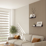 Modern living room with a light beige sofa, brown cushions, a potted plant, and sunlight streaming through striped blinds. Wooden slat accent wall features two black shelves with photos, a vase, and a decorative clock.