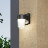 A modern wall-mounted outdoor light fixture with a black frame and clear glass shade is illuminated on a textured concrete wall, with greenery and a clear sky in the background.