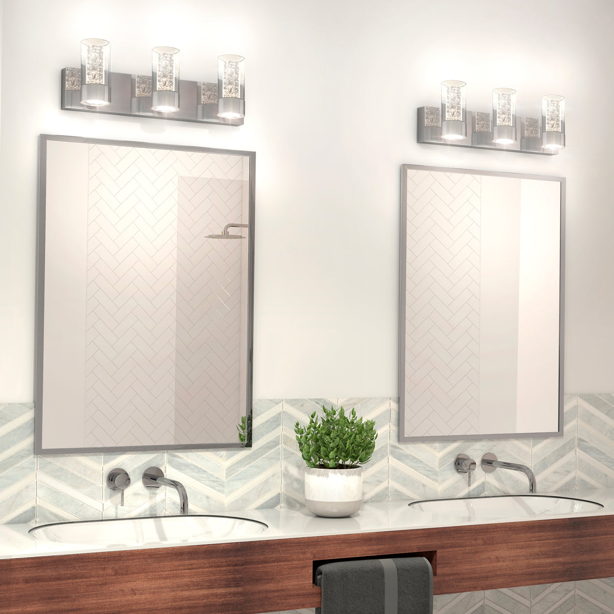 Modern bathroom with two rectangular mirrors above a wooden counter, each with an under-mount sink. A potted plant sits between the sinks, and wall-mounted lights are above each mirror. Chevron tile backsplash is visible.