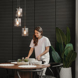 A woman in a white shirt arranges tableware on a round dining table with placemats and plants. Three pendant lights hang above, and a large potted plant stands beside the table against a dark, slatted wall.