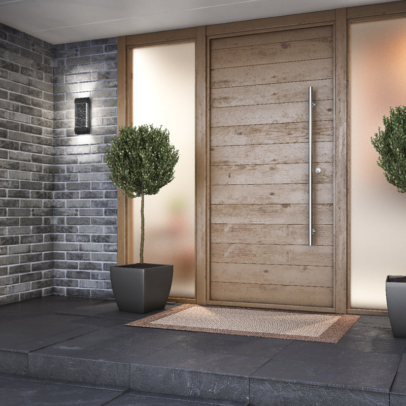 Modern wooden front door with a long vertical handle, frosted glass panels, two potted plants, a welcome mat, gray brick walls, and the Artika Essence Glow Box indoor-outdoor LED wall light.