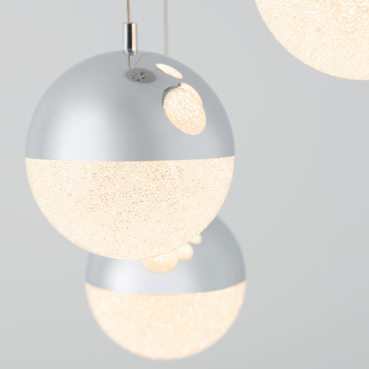 Close-up of modern spherical pendant lights with a metallic silver upper half and a textured, glowing white lower half, suspended against a plain light gray background.