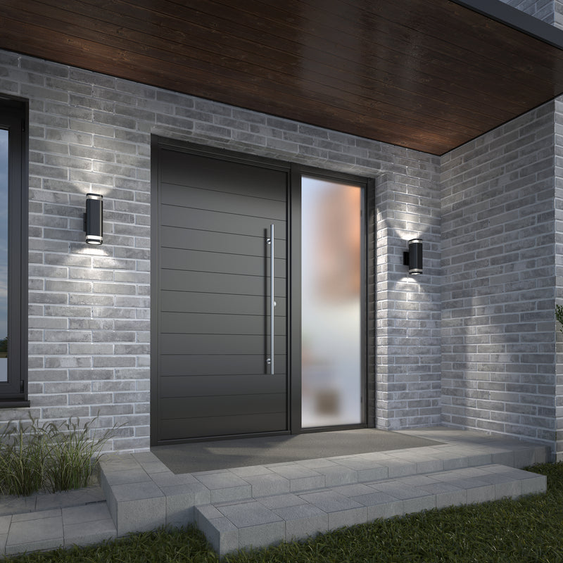 Modern front entrance with a large dark door, vertical handle, frosted glass side panel, gray brick walls, concrete step with grass at the side, and two Artika Mettle LED indoor-outdoor wall lights.