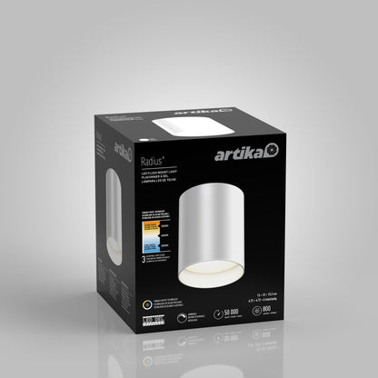 A matte black box for the Artika Radius LED flush mount light 3 CCT displays the cylindrical white fixture, highlights its adjustable color temperature, and includes details on brightness, lifespan, and energy efficiency.