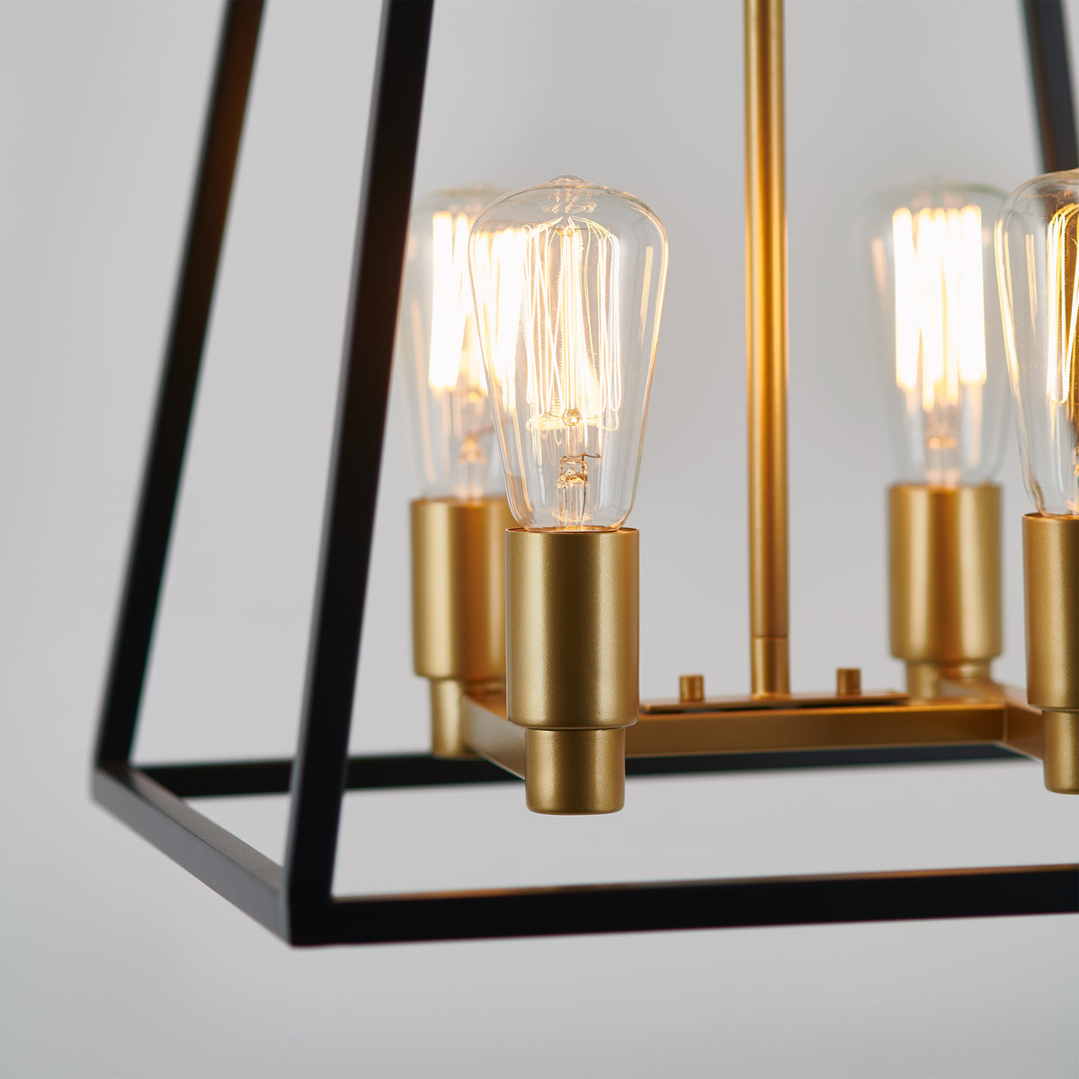 Close-up of a modern hanging light fixture with exposed Edison bulbs, gold sockets, and a black geometric metal frame against a plain light background.