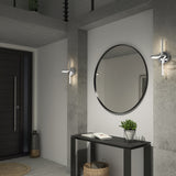 A modern entryway with concrete walls features a black console table, a round wall mirror, a potted plant, books, a basket underneath, two wall sconces, and a black front door in the background.
