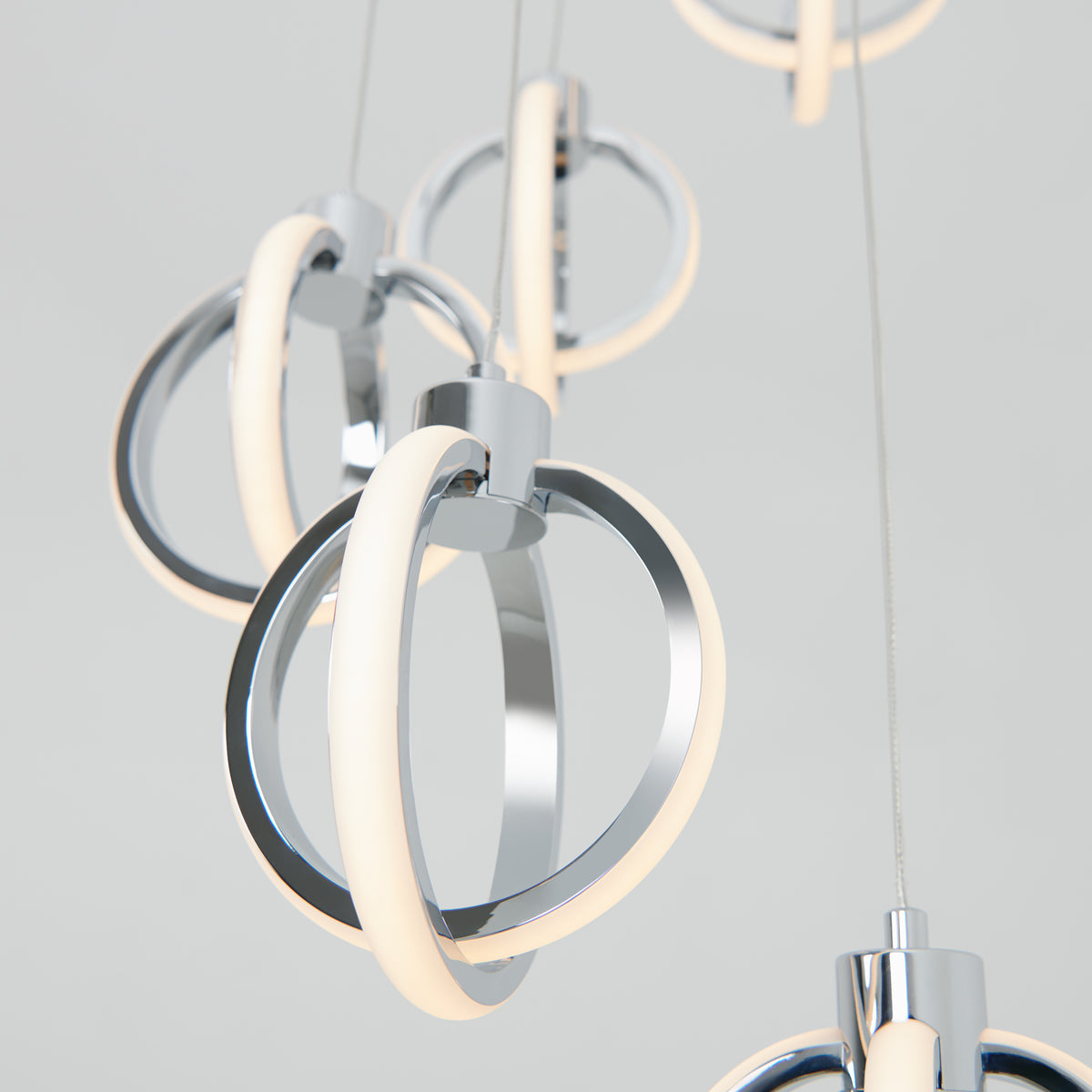 Close-up of modern hanging pendant lights with intertwined metallic rings and integrated LED light strips, suspended by thin cables against a neutral background.