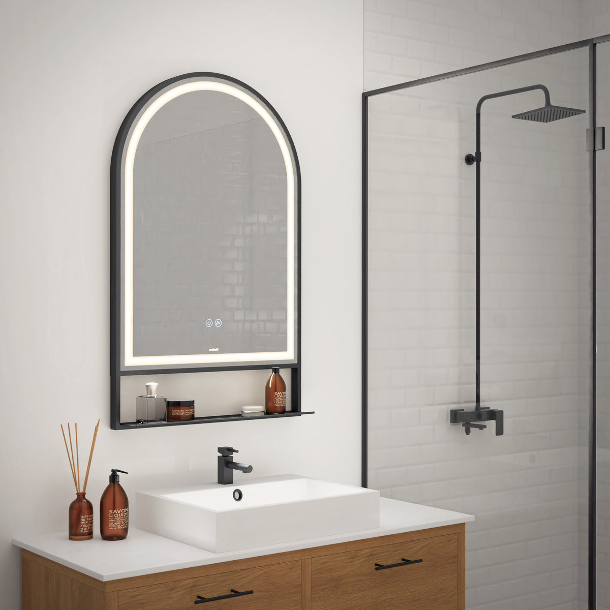 A modern bathroom with a wooden vanity, white sink, black faucet, and an arched LED mirror above. Glass shower enclosure with black fixtures is visible to the right. Brown bottles and a reed diffuser sit on the counter.